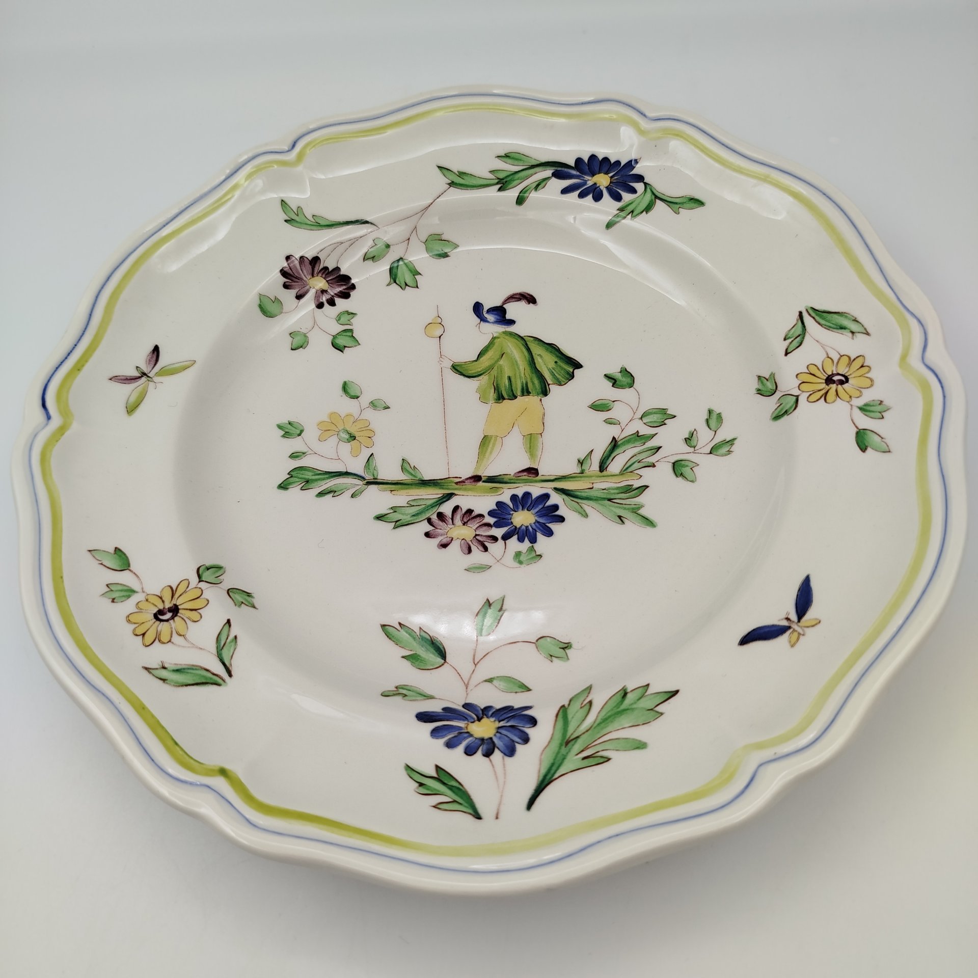 Тарелка обеденная, украшенная изображением цветов, бабочек и деревенского персонажа, узор "Moustiers", мануфактура "Longchamp", фаянс, деколь с подрисовкой, Франция, 1950-1970 гг.